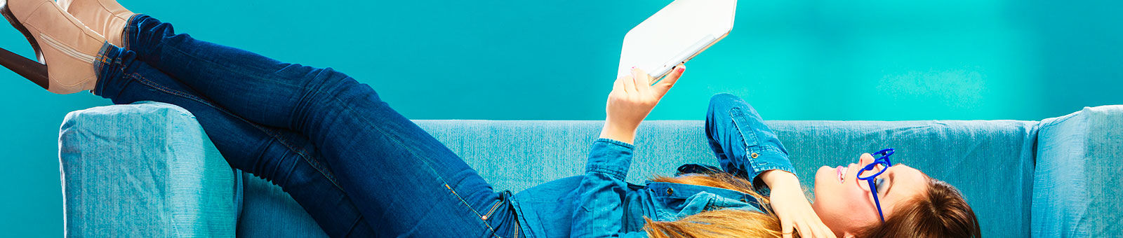 Jeune fille lisant via sa tablette numérique, alongée dans son canapé.