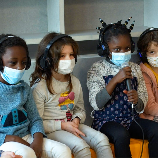 Une petite fille parle dans un micro assise à côté de ses camarades de classe